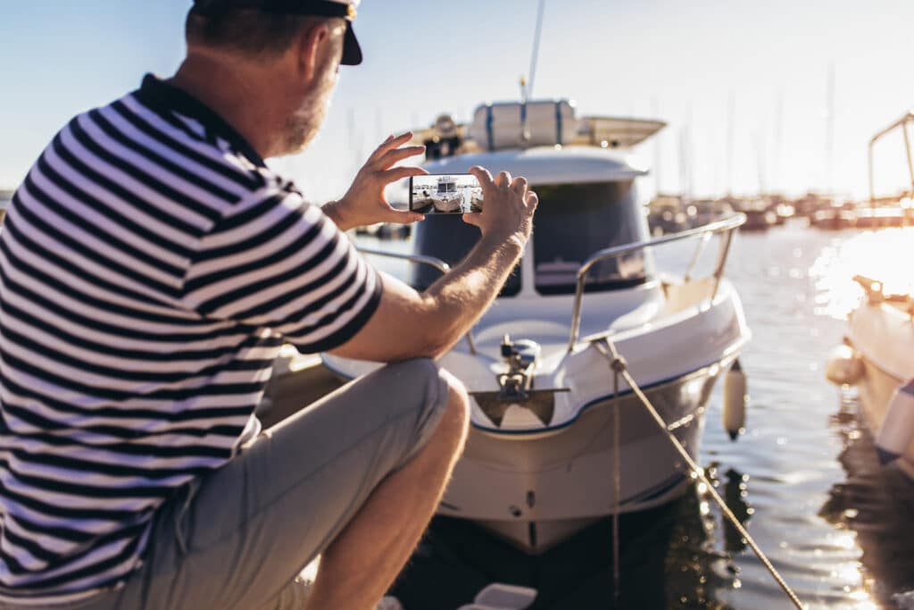 Mature man standing near the sea dressed in a sailor's shirt and hat taking a picture of his new boat with his smartphone